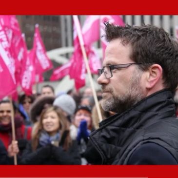 Man with glasses at a rally with pink flags.