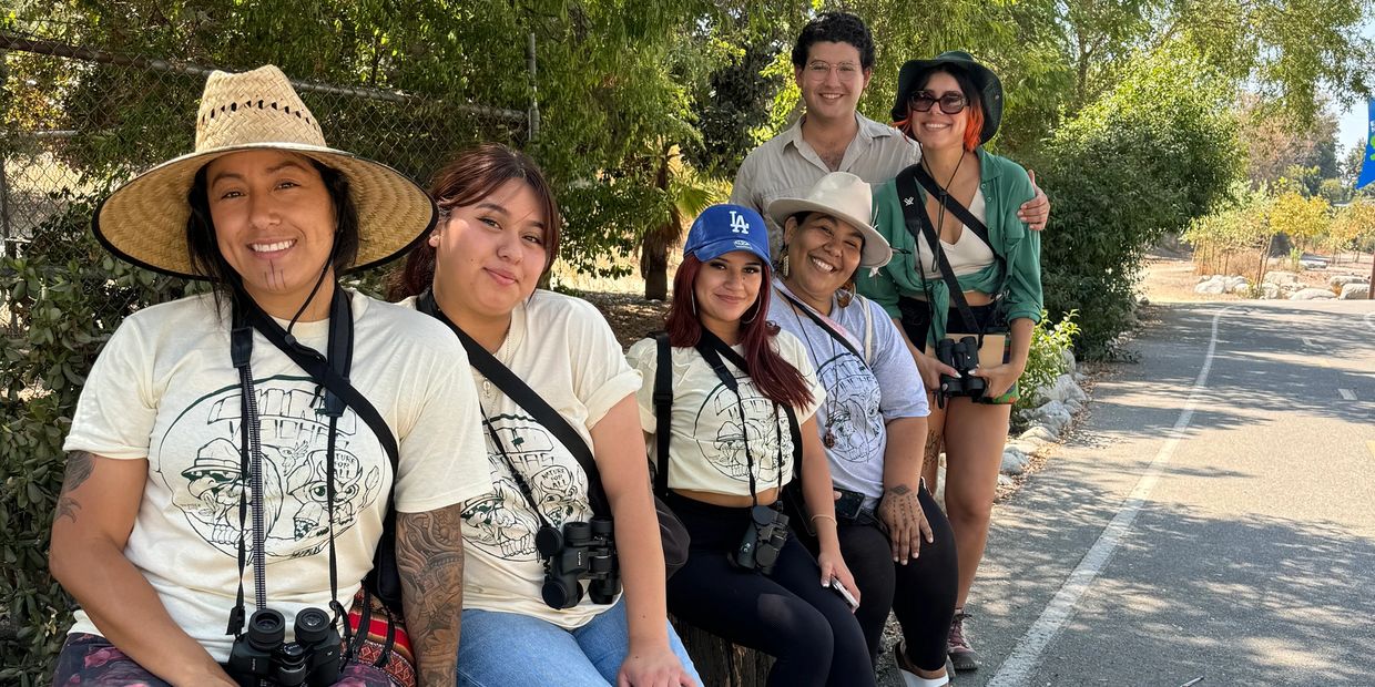 A group of six friends smiling outdoors with binoculars and hats, enjoying a sunny day.