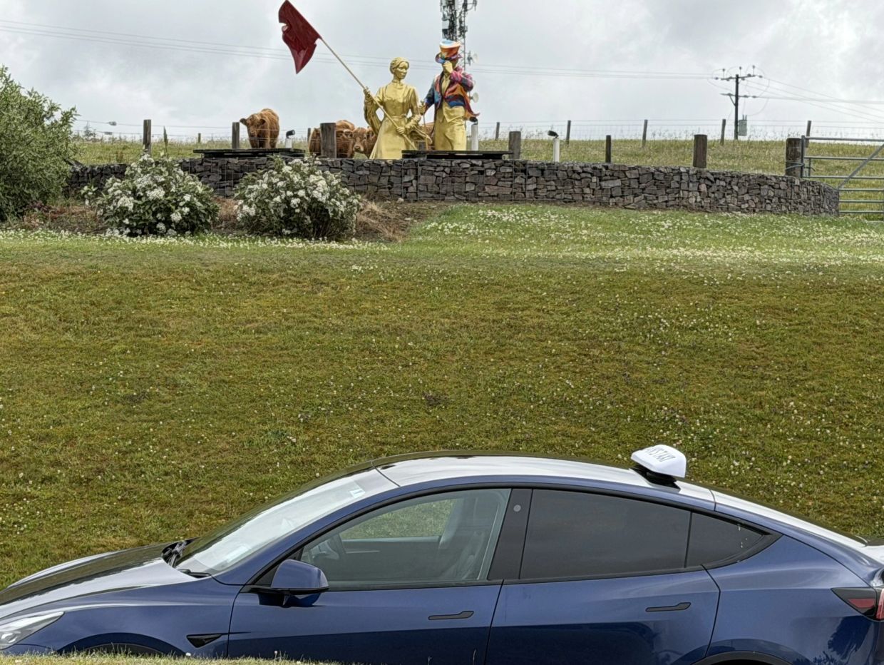 Blue car parked near a stone wall with statues and cows behind it.