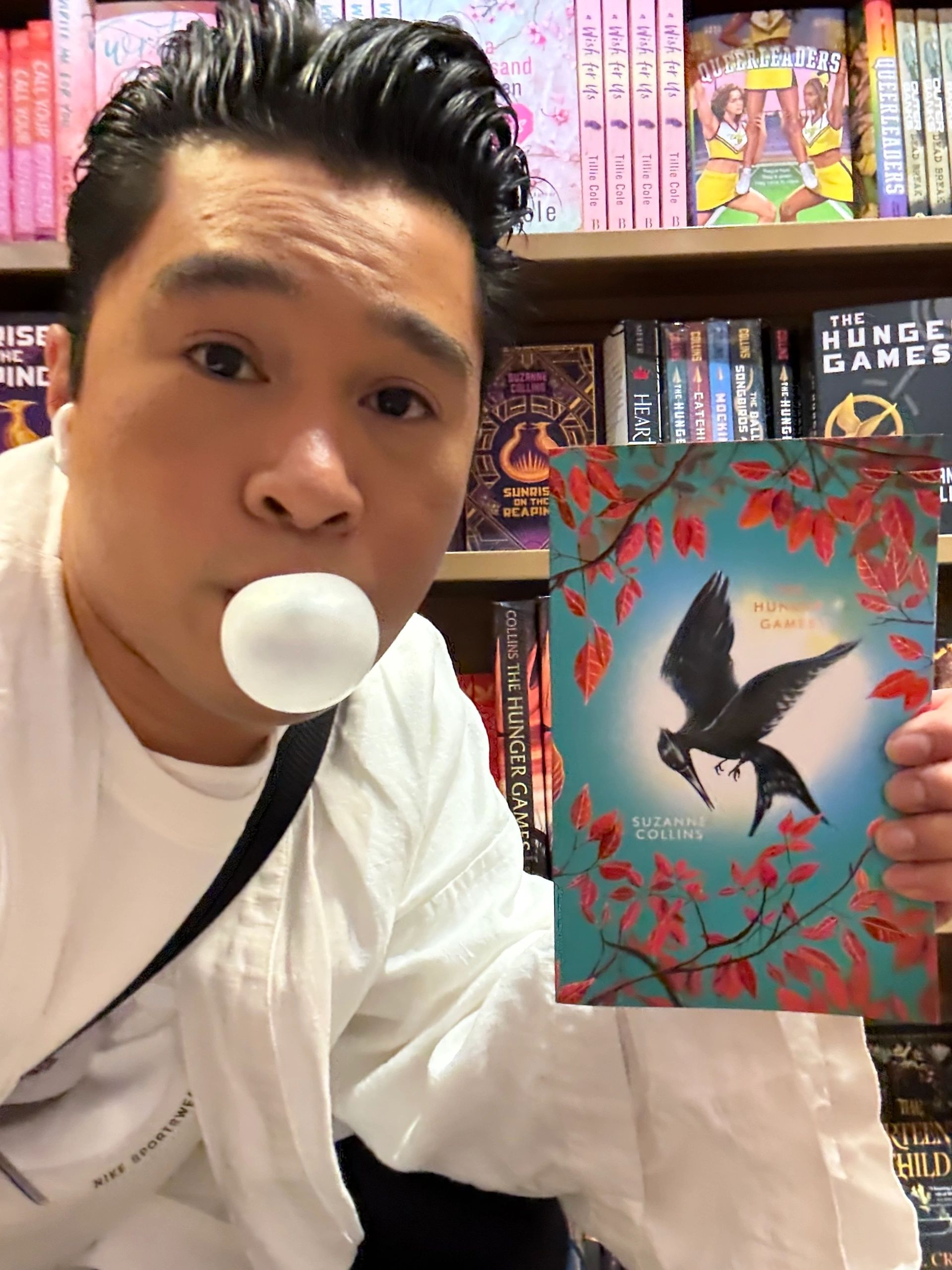 Person blowing bubble gum, holding The Hunger Games book in a bookstore.