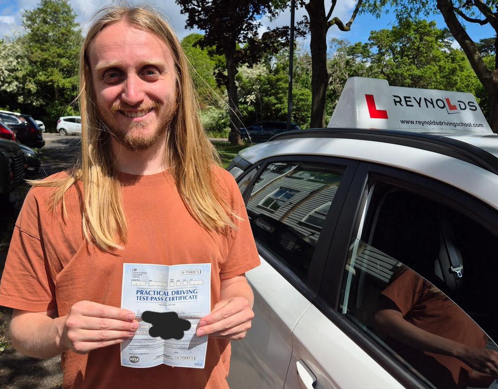 Man proudly holding a practical driving test pass certificate beside a driving school car.