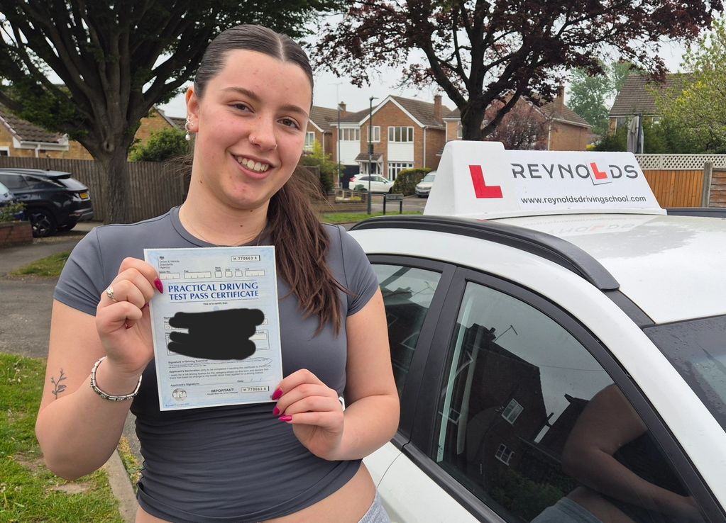 A smiling woman proudly holds her practical driving test pass certificate next to a learner driver car.