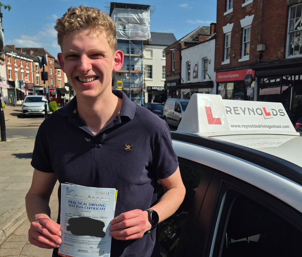 Young man smiling with his practical driving test pass certificate beside a driving school car.