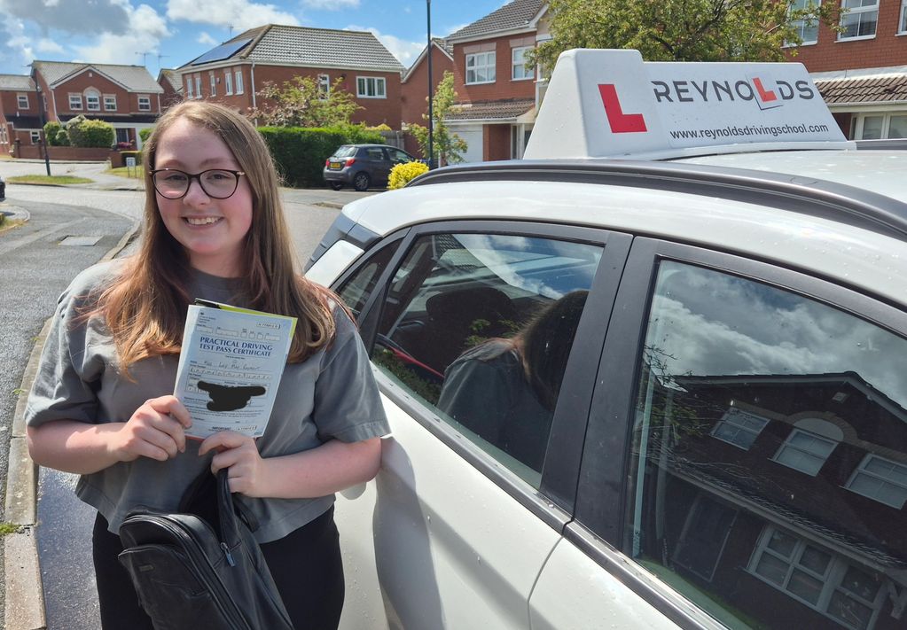 Young woman happily showing her driving test pass certificate beside a driving school car.