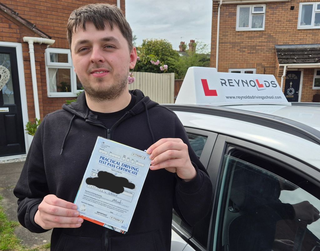 A man proudly holds his practical driving test pass certificate by a driving school car.