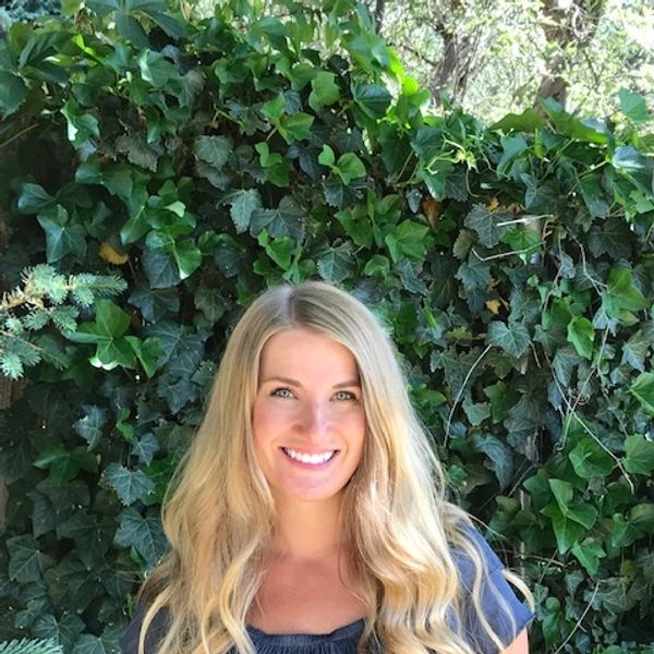 Smiling woman with long blonde hair stands before lush green ivy.