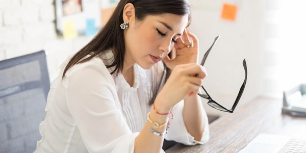 Woman holding glasses, looking stressed at a desk.