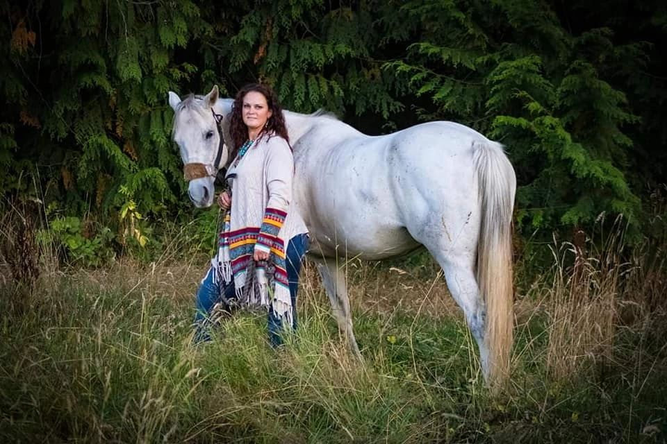 Woman standing in field with trees behind her next to grey quarter horse looking back over shoulder