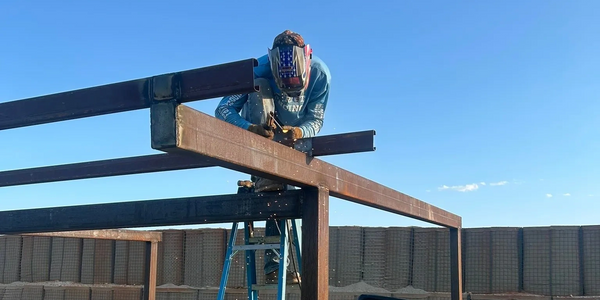 A worker welding metal beams on a ladder outdoors under clear skies.