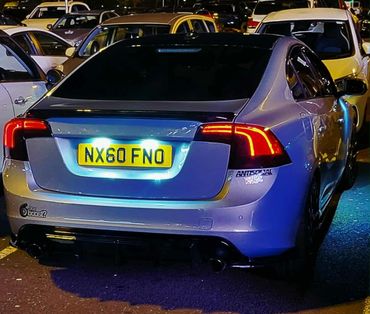 Silver car with illuminated license plate and custom stickers in a crowded parking lot at night.