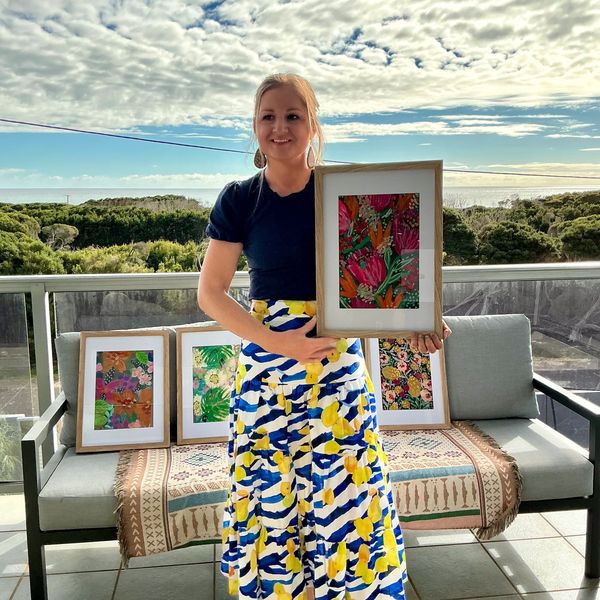 Sarah Hocking stands on a balcony, holding a bright and colourful protea painting. She is dressed in