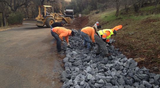 Paving crew lining ditch on right shoulder of community road with large rocks for erosion control