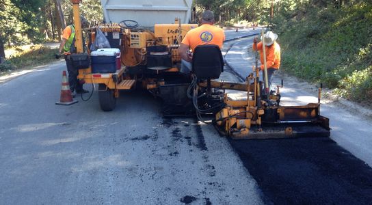 Paving crew filling narrow trench on right shoulder of community road with asphalt using machine.