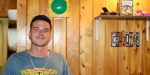 A young man in a blue Ace Hardware t-shirt stands against a wood-paneled wall with a traffic light and car-themed decor.