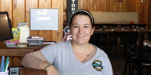 A smiling woman stands behind a wooden counter in a cozy restaurant with Route 66 decor.