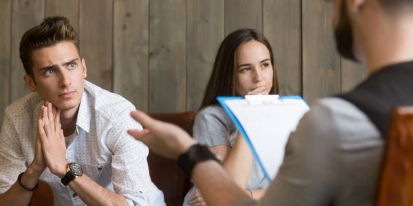 Frustrated young man listening to psychologist while his offended girlfriend sitting apart