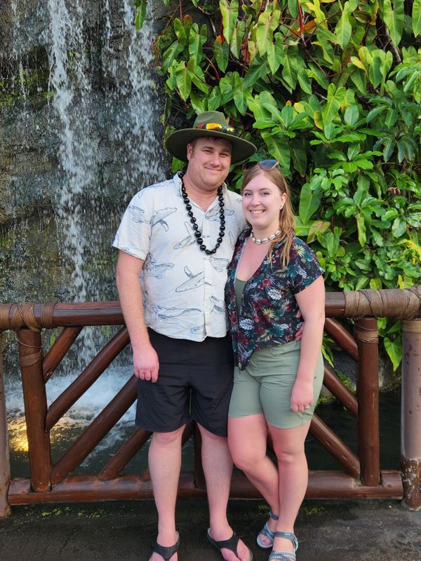 Couple posing happily by a wooden railing with a waterfall and lush greenery behind them.