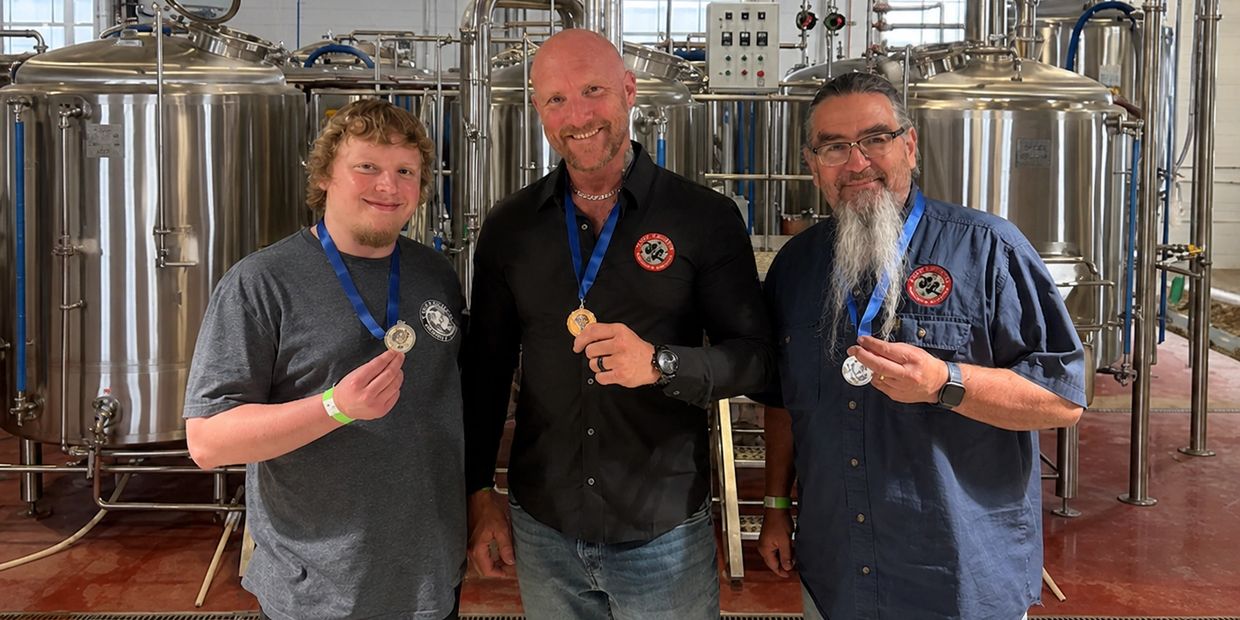 Three men proudly display medals in a brewery setting.