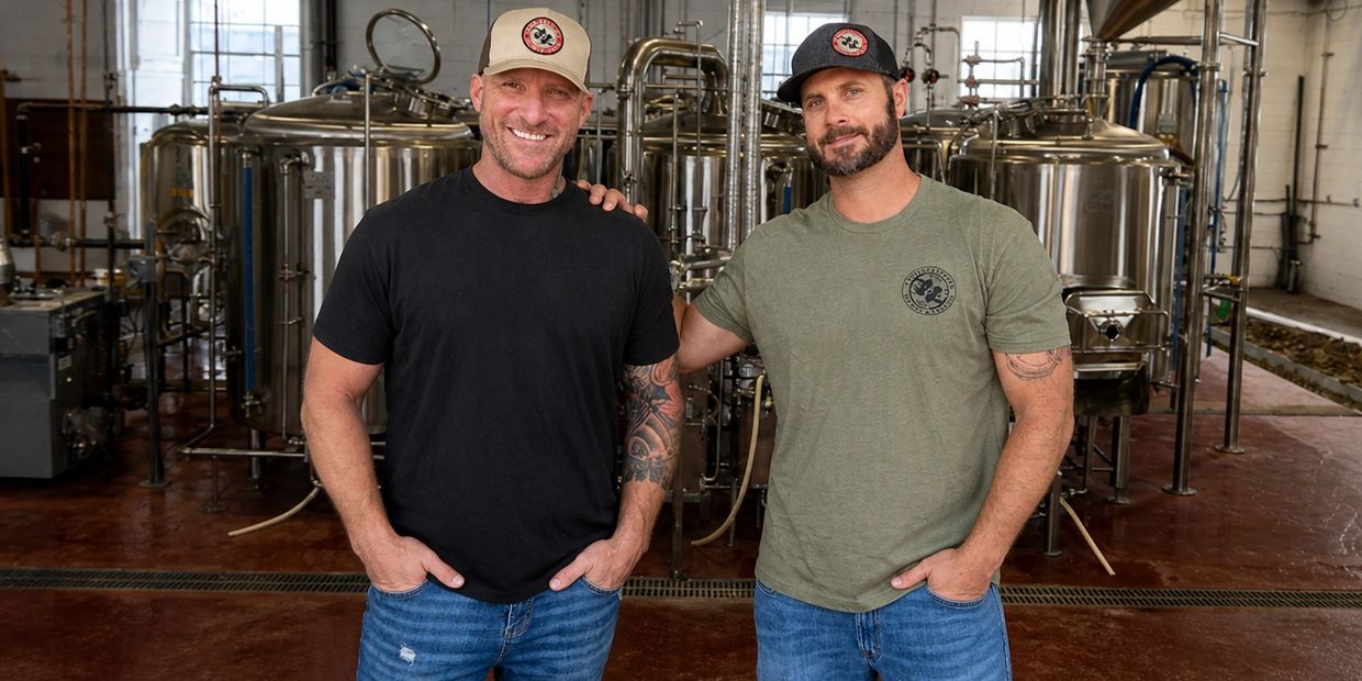 Two men stand smiling in a brewery with stainless steel tanks behind them.