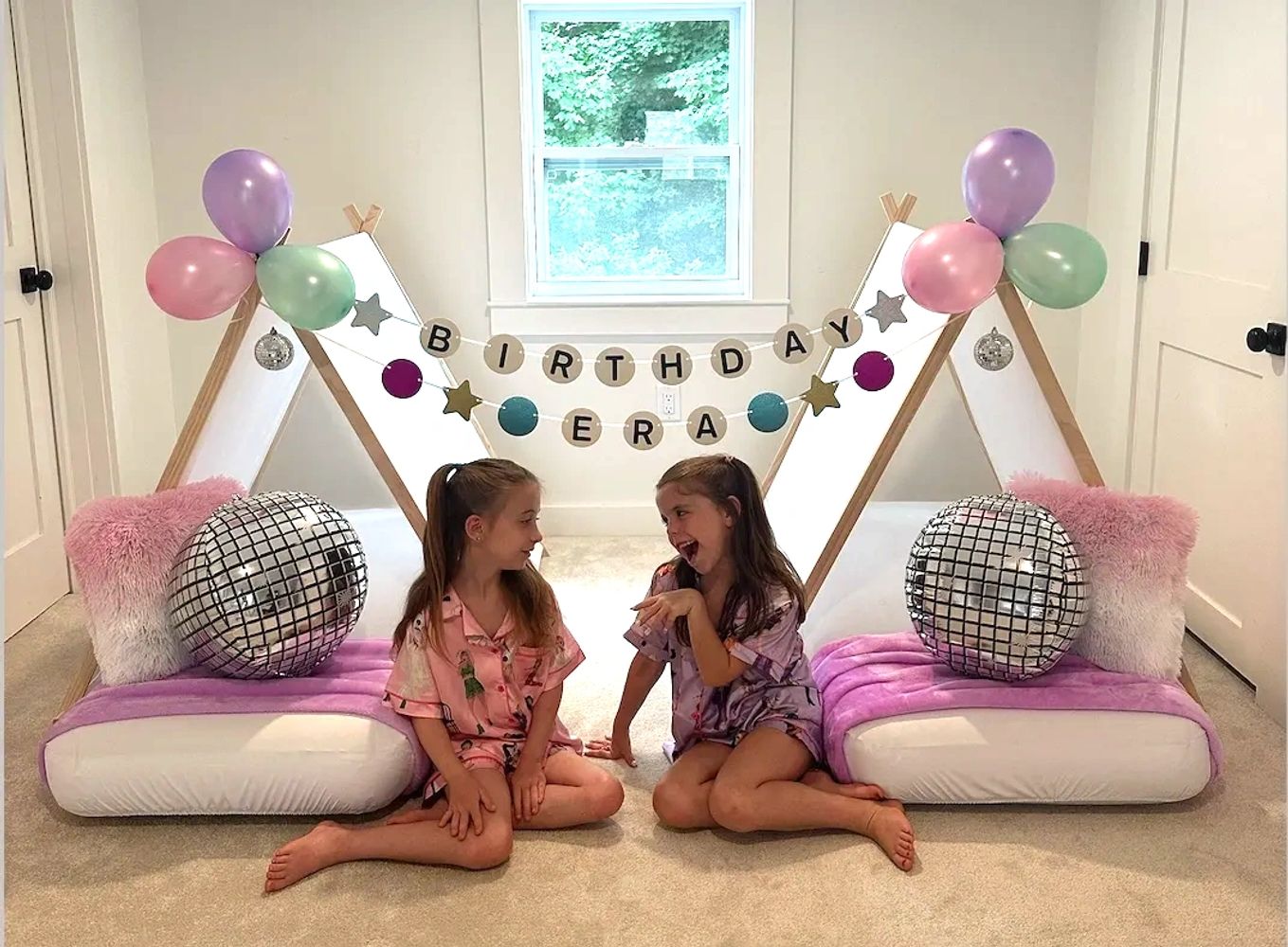 Two girls in pajamas sitting by decorated tents for a birthday party.