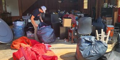 Person organizing a cluttered garage filled with bags and furniture.