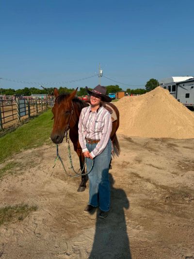 A woman in cowboy attire stands with a brown horse.