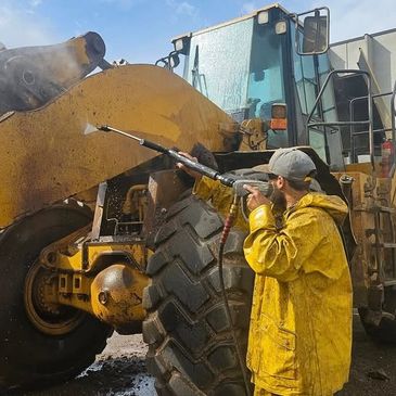 Worker in yellow raincoat pressure washes a large industrial vehicle outdoors.