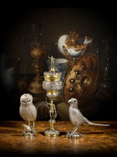 Intricately crafted silver bird sculptures on a wooden surface with antique vessels in the background.