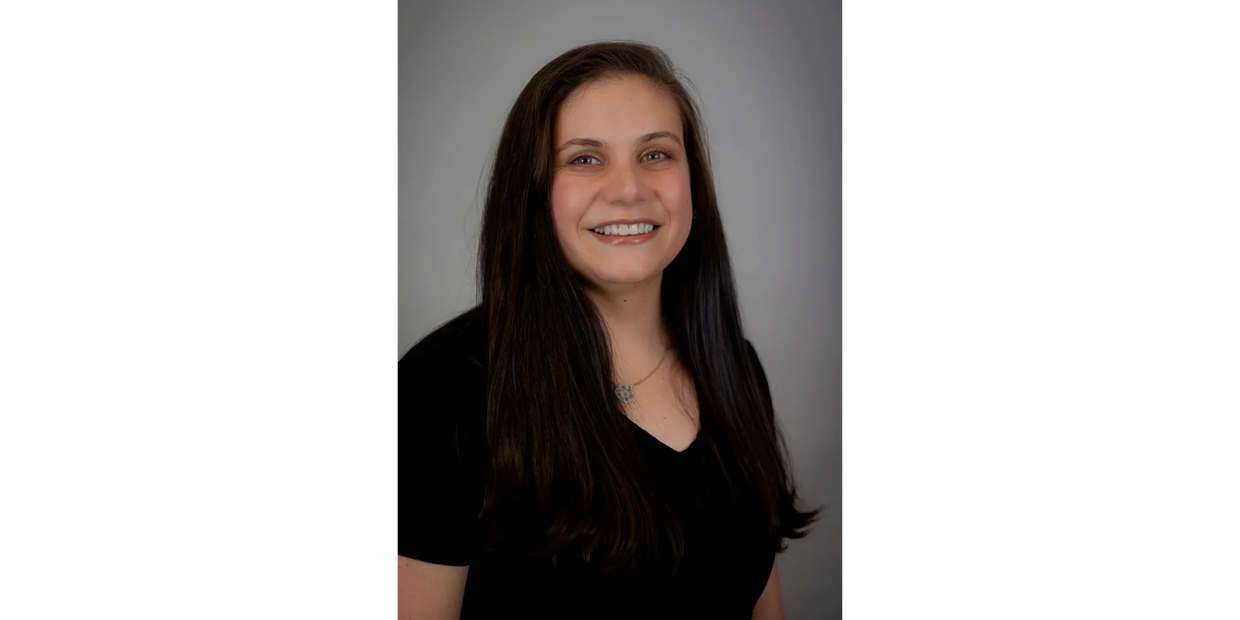 Smiling young woman with long dark hair in a black top against a neutral gray background.