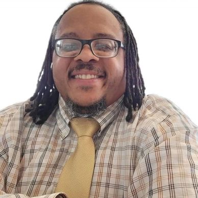 Smiling man wearing glasses, a plaid shirt, and a yellow tie.