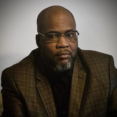 A serious man wearing a brown plaid blazer and glasses.