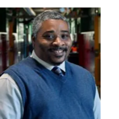 Smiling man in a blue sweater vest in a hallway.