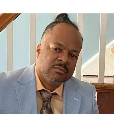 Man in a light blue suit and patterned tie sitting by a staircase.
