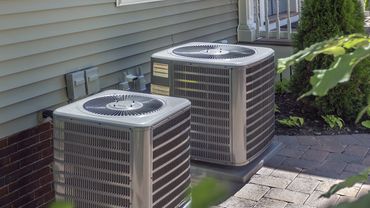 Two outdoor air conditioning units beside a house on a paved patio.