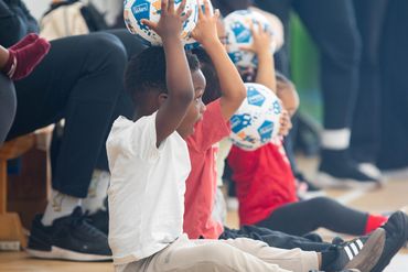 Children sitting on the floor holding balls above their heads indoors.