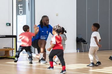 A coach interacts with young children in a sports hall, encouraging and playing.