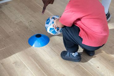 Child holding a soccer ball next to a blue training cone indoors.