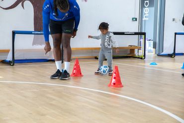 Child learns soccer dribbling with adult coach indoors.