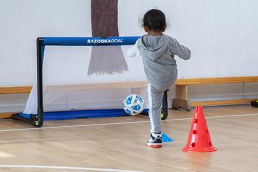 A child kicks a soccer ball towards a small goal during indoor practice.