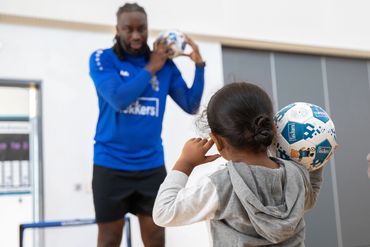 A coach teaches children how to throw a soccer ball indoors.