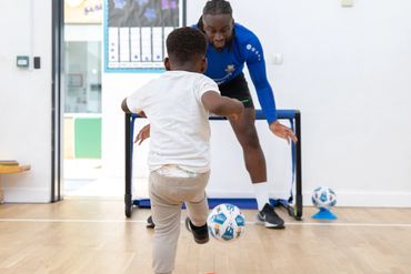 A child kicks a soccer ball towards a man acting as goalie indoors.