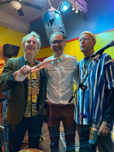 Three men posing together in a colorful room, one holding a wooden paddle.