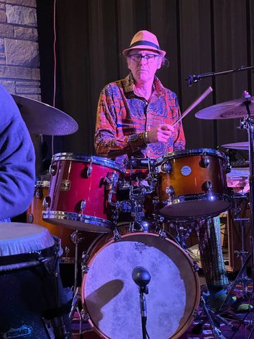 A drummer in a patterned shirt and hat playing on a drum set under stage lights.