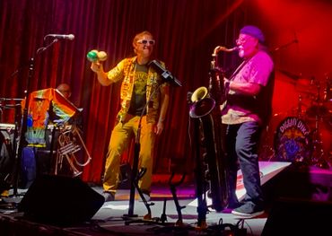 Two musicians perform energetically on stage with vibrant lighting and colorful outfits.