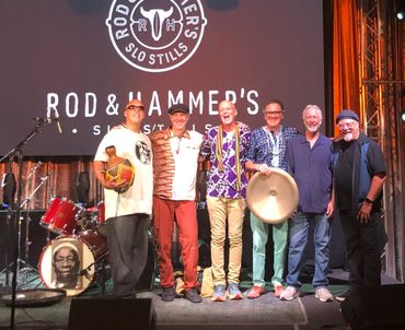 A joyful group of six musicians posing on stage with instruments and a Rod & Hammer's backdrop.