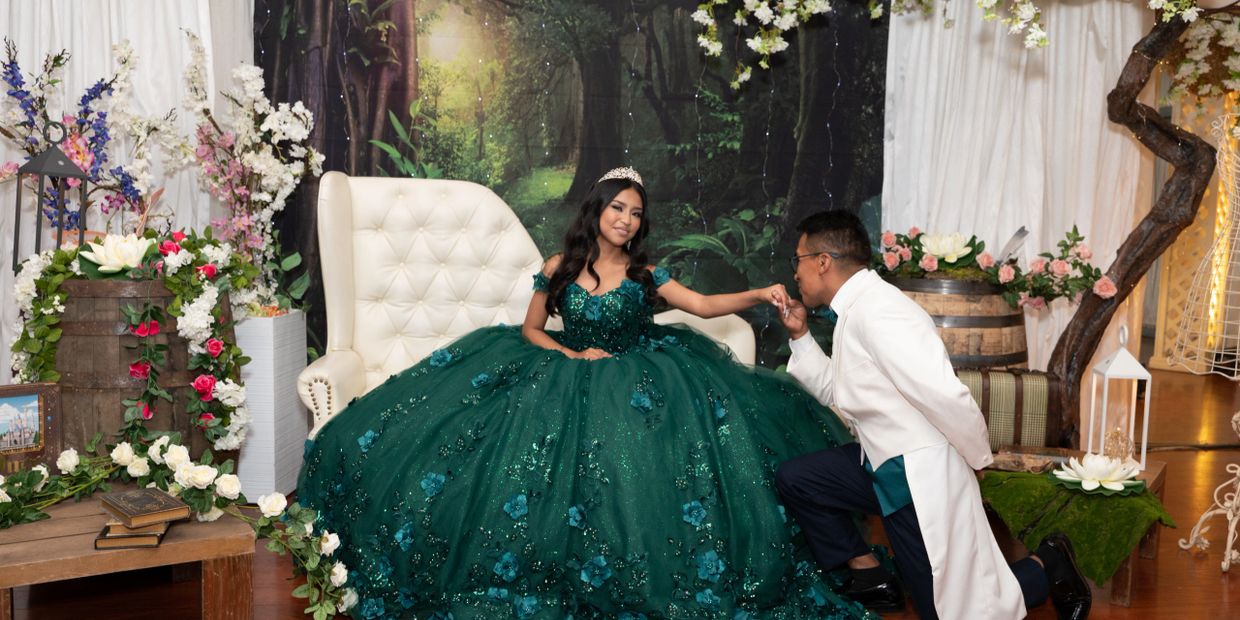 A young woman in a green gown with a tiara, a man kneeling and kissing her hand in a floral setting.