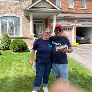 Two people smiling outside a suburban house on a sunny day.