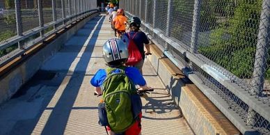 Group of young kids wearing bike helmets,  riding bicycles single-file across a bridge.