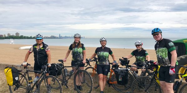 Five cyclists standing with bikes along Lake Michigan. Chicago skyline in the distance.