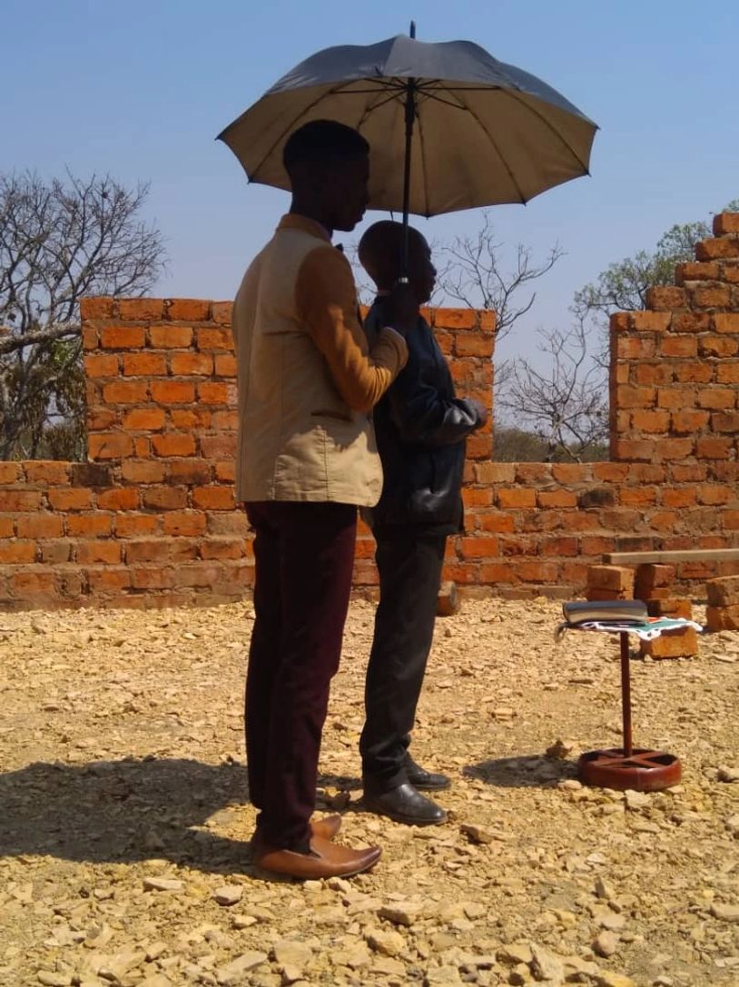 Two men standing under an umbrella near a brick wall.